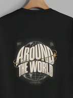 A black short-sleeve t-shirt hanging on a wooden hanger against a solid gray background. The back of the shirt features a large graphic print with the words 'AROUND THE WORLD' in a stylized white and gold font, surrounding a globe illustration.
