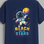 A navy blue short-sleeve t-shirt hanging on a wooden hanger against a solid gray background. The back of the shirt features a colorful graphic of an astronaut floating in space, reaching toward a yellow and purple planet, with the text 'REACH FOR THE STARS' written below in yellow and blue.