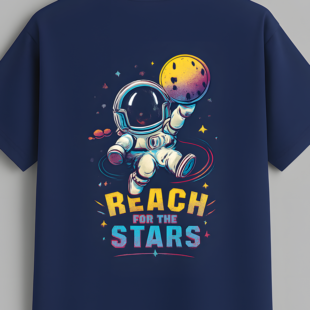 A navy blue short-sleeve t-shirt hanging on a wooden hanger against a solid gray background. The back of the shirt features a colorful graphic of an astronaut floating in space, reaching toward a yellow and purple planet, with the text 'REACH FOR THE STARS' written below in yellow and blue.