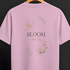 A light pink short-sleeve t-shirt hanging on a wooden hanger against a dark gray background. The back of the shirt features a graphic print with the text 'BLOOM OWN TIME' in black and gold, surrounded by delicate gold and green floral illustrations.