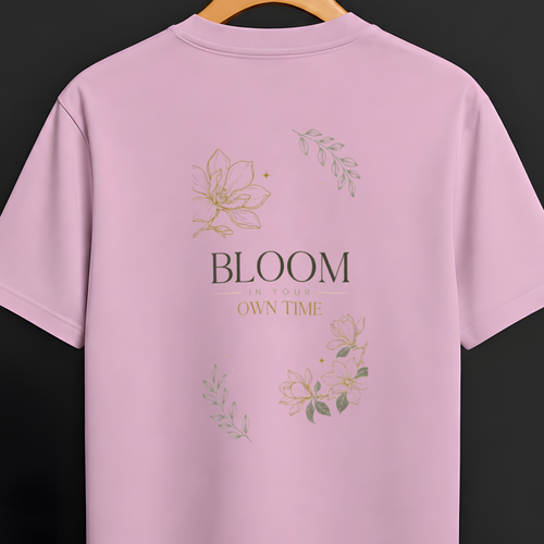 A light pink short-sleeve t-shirt hanging on a wooden hanger against a dark gray background. The back of the shirt features a graphic print with the text 'BLOOM OWN TIME' in black and gold, surrounded by delicate gold and green floral illustrations.