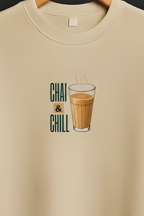 A beige short-sleeve crewneck t-shirt hanging on a wooden hanger against a dark gray background. The shirt features a graphic on the chest with the text 'CHAI & CHILL' in green and a brown iced coffee cup illustration.