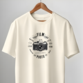 An off-white short-sleeve t-shirt with a black vintage film camera graphic and circular text on the chest, hanging on a wooden hanger from a clothing rack in front of a gray wall and white door.