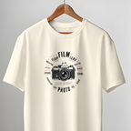 An off-white short-sleeve t-shirt with a black vintage film camera graphic and circular text on the chest, hanging on a wooden hanger from a clothing rack in front of a gray wall and white door.