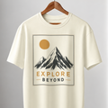 An off-white short-sleeve crewneck t-shirt hanging on a wooden hanger from a clothing rack. The shirt features a graphic print of a mountain under a yellow sun with the text 'EXPLORE BEYOND'.