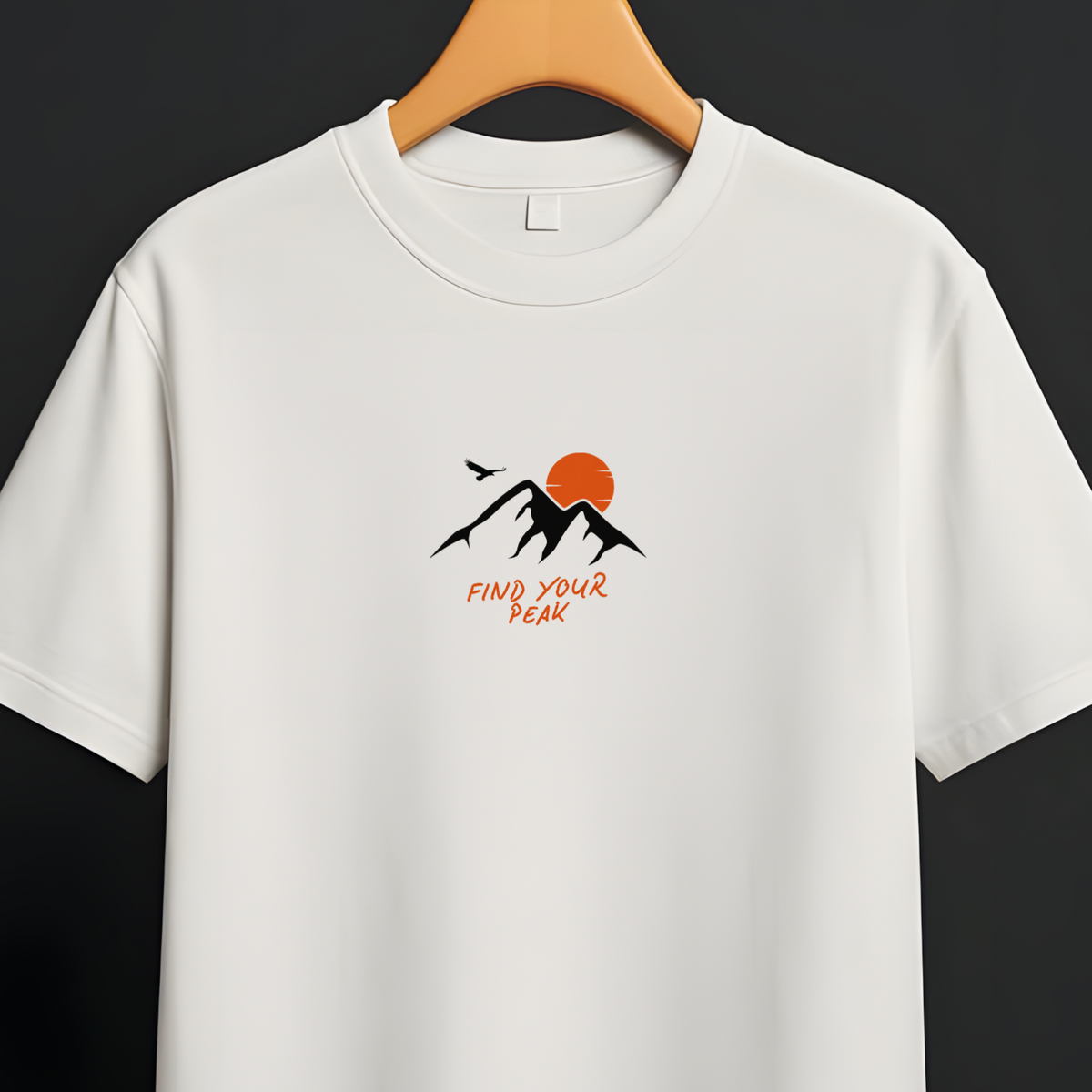 A white short-sleeve crewneck t-shirt hanging on a wooden hanger from a black clothing rack against a dark gray background. The shirt features a small graphic on the center chest with black mountains, an orange sun, and orange text reading 'find your peak'.