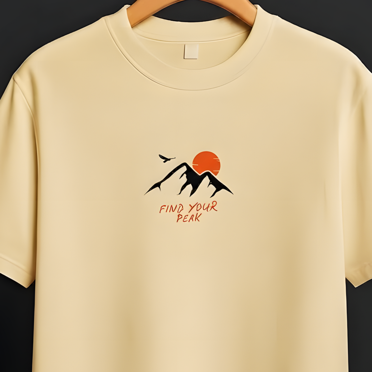 A cream-colored short-sleeve t-shirt hanging on a wooden hanger against a dark gray background. The shirt features a small graphic on the left chest with black mountains, an orange sun, a bird, and the text 'PUSH YOUR KIS'.