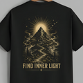 A black short-sleeve t-shirt hanging on a wooden hanger against a solid gray background. The back of the shirt features a large, detailed graphic print in a warm beige or gold tone, showing a mountain peak with a glowing sunburst, swirling mist, a winding path, and the text 'FIND INNER LIGHT' printed below.