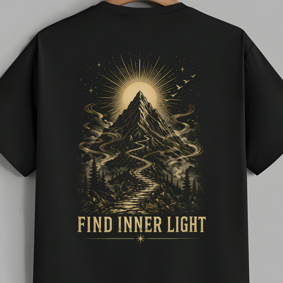 A black short-sleeve t-shirt hanging on a wooden hanger against a solid gray background. The back of the shirt features a large, detailed graphic print in a warm beige or gold tone, showing a mountain peak with a glowing sunburst, swirling mist, a winding path, and the text 'FIND INNER LIGHT' printed below.