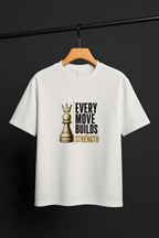 A white short-sleeve crewneck t-shirt hanging on a wooden hanger from a black clothing rack. The shirt features a graphic print of a gold chess queen piece next to the text 'EVERY MOVE BUILDS STRENGTH' in black and gold.