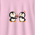 A light pink short-sleeve crewneck t-shirt hanging on a wooden hanger against a neutral gray background. The front of the shirt features a cute graphic of two cartoon penguins sharing a small cake with hearts and sparkles.