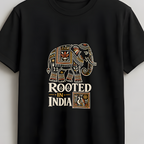 A black short-sleeve t-shirt hanging on a wooden hanger against a light gray wall. The shirt features a graphic print of an ornate elephant in cream, gold, and brown, with the text 'ROOTED IN INDIA' underneath.