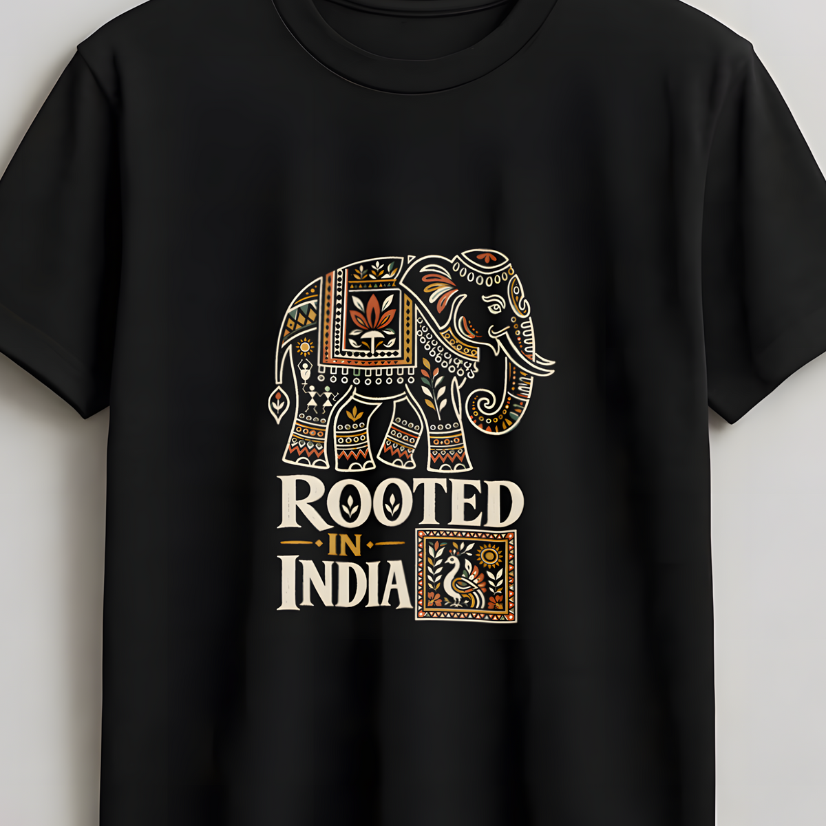 A black short-sleeve t-shirt hanging on a wooden hanger against a light gray wall. The shirt features a graphic print of an ornate elephant in cream, gold, and brown, with the text 'ROOTED IN INDIA' underneath.
