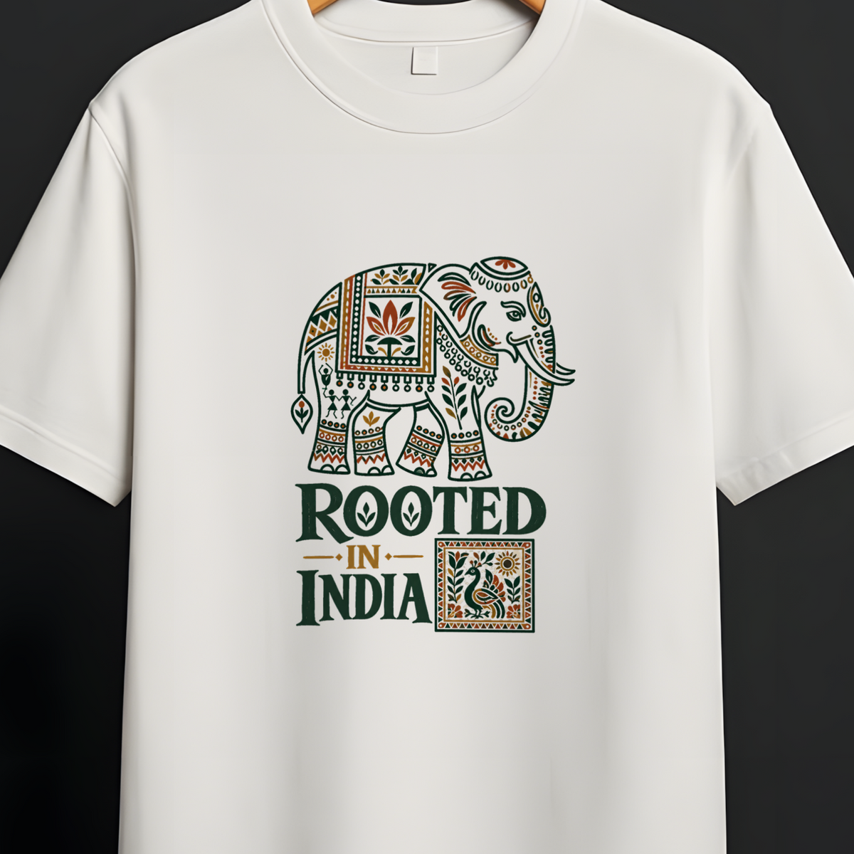 A white short-sleeve crewneck t-shirt hanging on a wooden hanger against a dark gray background. The shirt features a graphic print of an ornate elephant and the text 'ROOTED IN INDIA' in dark green and gold.