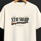 An off-white short-sleeve t-shirt hanging on a wooden hanger against a dark background. The shirt features a graphic print on the front with the text 'STAY SHARP' and a vintage-style rifle illustration.