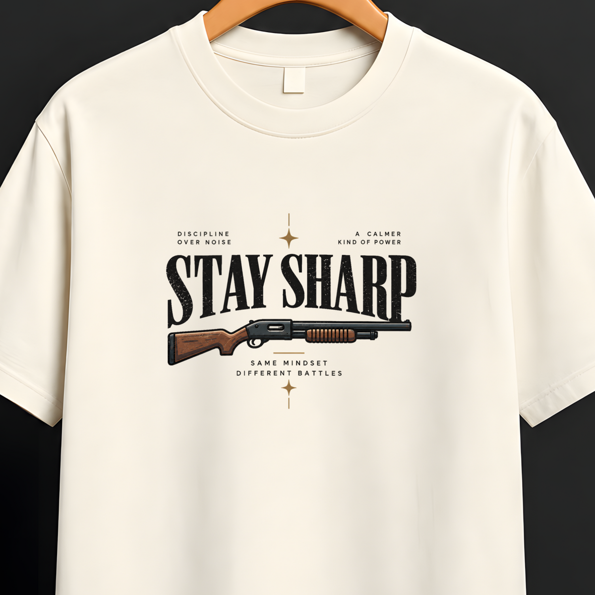 An off-white short-sleeve t-shirt hanging on a wooden hanger against a dark background. The shirt features a graphic print on the front with the text 'STAY SHARP' and a vintage-style rifle illustration.